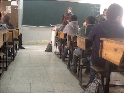 Photo of children sitting at desks in a classroom with teacher standing at the front. Some chalk writing in Arabic on the blackboard.