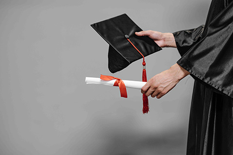 a partial image of a person in a graduation gown, holding out their graduation cap and scroll