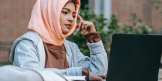 A student wearing a headscarf sits outside in front of a laptop computer with one hand pointed to her head in contemplation, and one hand on the laptop